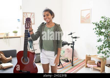 Fröhliches Schulmädchen mit Gitarre, das in der Mitte des häuslichen Raumes gegen das Schlagzeug steht und dich ansieht Stockfoto