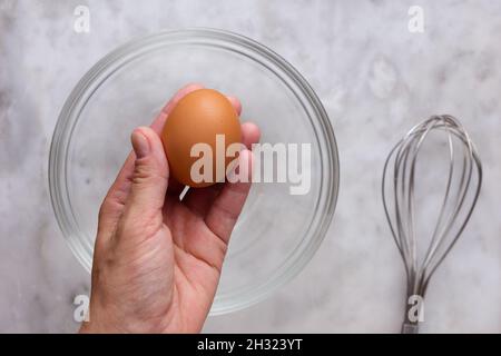 Draufsicht auf die linke Frauenhand, die ein rohes braunes Ei über der Glasschale hält und auf der Marmoroberfläche Schneebesen Stockfoto