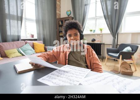 Happy cute afrikanischen Schuljunge mit Papieren sitzen am Tisch während der Lektion in häuslicher Umgebung Stockfoto