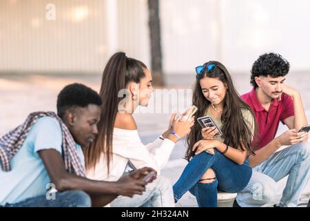 Junge Menschen unterschiedlicher Ethnien sitzen draußen und schauen sich ihre Handys an Stockfoto