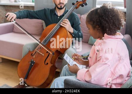 Interkulturelles Mädchen, das Musiknoten auf Papier schreibt, während es vor ihrer Musiklehrerin sitzt und Cello spielt Stockfoto