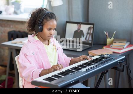 Hübsches Schulmädchen in Kopfhörern, das Klaviertastatur spielt, während es während des Online-Unterrichts den Ratschlägen ihrer Lehrerin zuhört Stockfoto