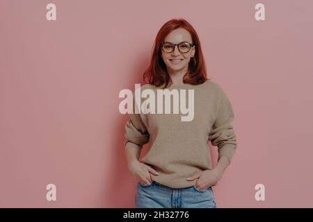 Glückliche rothaarige Schülerin in einer Brille lächelt an der Kamera, isoliert auf rosa Hintergrund Stockfoto