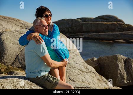 Liebevolles reifes Paar auf Reisen, auf dem Felsen sitzend, erkunden. Echter Mann und Frau umarmen, küssen und lächeln glücklich. Skandinavische Landschaft, Norwegen Stockfoto