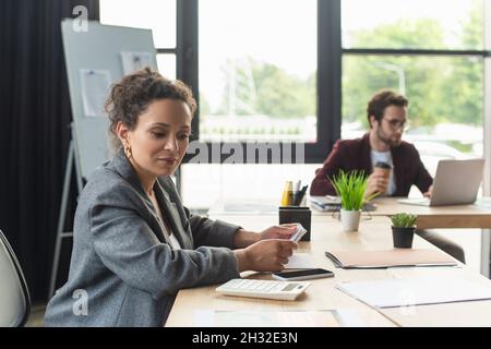 afroamerikanische Geschäftsfrau, die den Rechner in der Nähe des Smartphones und der Dokumente im Büro ansieht Stockfoto