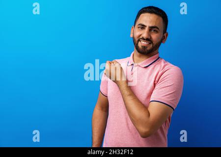Hispanic Mann mit Bart trägt lässiges T-Shirt zeigt Arme Muskeln vor blauem Hintergrund Stockfoto