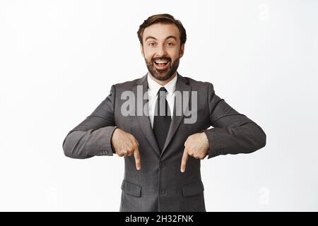 Der enthusiastische Geschäftsmann im Anzug zeigt die Finger nach unten, lächelt und zeigt Werbung, zeigt Diagramm oder Diagramm unten, weißer Hintergrund Stockfoto
