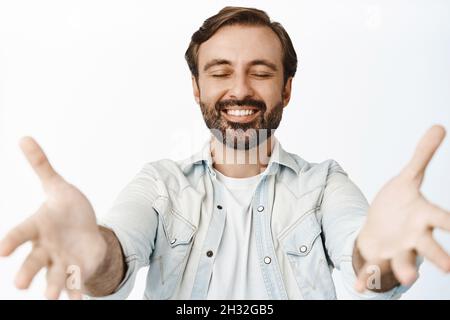 Lächelnder bärtiger Kerl schließt die Augen, greift nach etwas, empfängt in Händen, steht über weißem Hintergrund Stockfoto