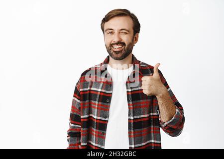 Gutaussehender lächelnder Familienmensch zwinkerte und zeigte Daumen nach oben, empfahl Produkt, Lob und mag Promo-Angebot, stand auf weißem Hintergrund Stockfoto