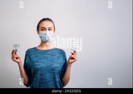 Medizinische Wahl. Junge Frau in einer medizinischen Maske auf weißem Hintergrund hält Blasen von Pillen aus dem Virus. Blick direkt auf die Kamera. Epidemie Stockfoto