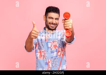 Glücklicher, zufriedener Mann mit Bart und dunklem Haar, das rotes Retro-Telefon aushält und den Daumen vor die Kamera zeigt, ein blaues Hemd trägt und mit toothy Lächeln posiert. Innenaufnahme des Studios isoliert auf rosa Hintergrund. Stockfoto