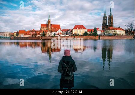 Frau in Winterkleidung blickt auf eine europäische Innenstadt auf der anderen Seite des Sees. Rückansicht einer Frau, die die Stadt mit einem roten Dach ansah, Abendansicht Stockfoto