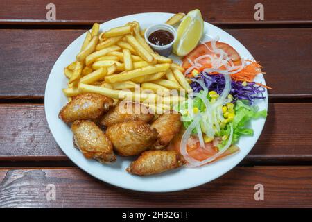 Gegrillte Hähnchenschenkel in Sauce und garniert mit Bratkartoffeln und veganem Salat. Auf einem hölzernen Hintergrund, Nahaufnahme. Stockfoto