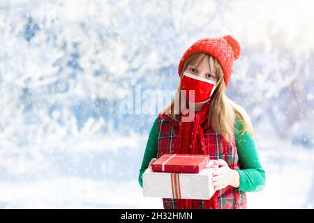 Kind in Gesichtsmaske mit Weihnachtsgeschenk. Kind trägt schützende medizinische Maske für sichere Weihnachtsfeier. Familienbesuch bei Ausbruch von Covid-19. Stockfoto