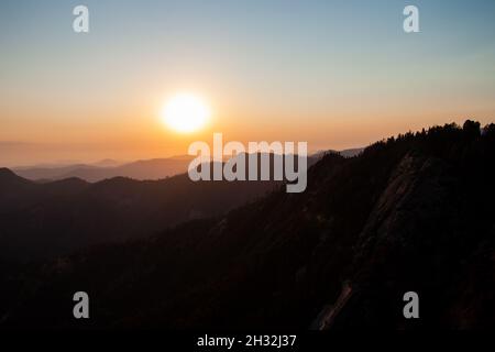 Wunderschöner Sonnenuntergang über den Bergen, die von Wald überwuchert sind, riesige orangefarbene Sonne geht unter, blauer Himmel | szenischer Sonnenuntergang am Moro Rock im Sequoia National Park USA Stockfoto