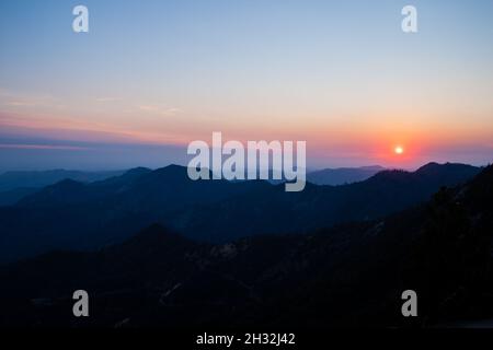 Farbenprächtiger Sonnenuntergang über den Bergen mit Wald überwuchert, orangefarbene Sonne geht unter, rosa-blauer Himmel | Szenischer Sonnenuntergang am Moro Rock im Sequoia National Park USA Stockfoto