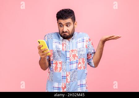 Porträt von attraktiven bärtigen Mann trägt blaue Casual Shirt halten Smartphone in den Händen, Verbreitung hans beiseite, nicht wissen, wie Anwendung zu verwenden. Innenaufnahme des Studios isoliert auf rosa Hintergrund. Stockfoto