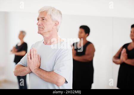 Älterer Mann steht mit verbundenen Händen und meditiert während des Yoga-Übens im Studio Stockfoto