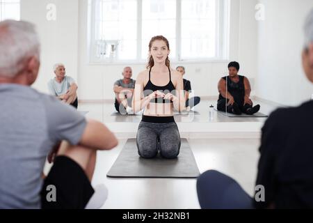 Junge, gesunde Frau, die während des Yoga-Kurses im Studio auf der Trainingsmatte sitzt und mit älteren Menschen spricht Stockfoto