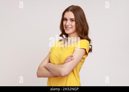 Portrait einer positiven jungen Frau mit braunem welligen Haar in gelbem T-Shirt, das mit gekreuzten Armen steht und mit Freundlichkeit und einem charmanten Lächeln auf die Kamera blickt. Innenaufnahme des Studios isoliert auf grauem Hintergrund. Stockfoto