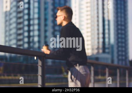 Ein nicht erkennbarer junger Mann im Business-Anzug lehnt sich an einem Metallgeländer an einem Pier gegen mehrstöckige Wohngebäude. Nachdenklich, reflektierend, in Stockfoto