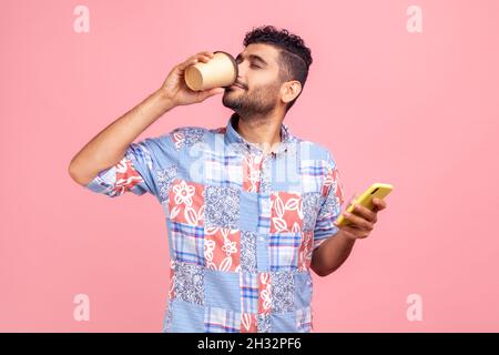 Portrait von attraktiven jungen erwachsenen Mann in lässigem Stil Hemd stehend und hält Einweg-Tasse und Telefon, trinken und genießen heiße Getränke. Innenaufnahme des Studios isoliert auf rosa Hintergrund. Stockfoto