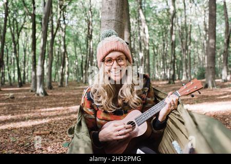 Frau lacht, während sie die Ukulele auf einer Hängematte im Wald spielt Stockfoto