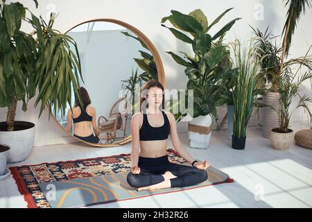 Junge Frau in Sportkleidung praktiziert Lotusstellung zu Hause Stockfoto