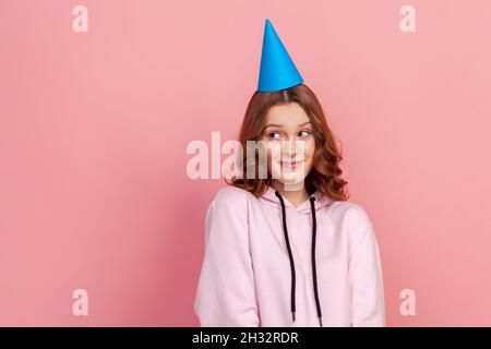 Porträt von schüchternen lockigen Teenager-Mädchen in Hoodie und blauen Party Hut suchen nach oben, Geburtstagsgrüße. Innenaufnahme des Studios isoliert auf rosa Hintergrund Stockfoto