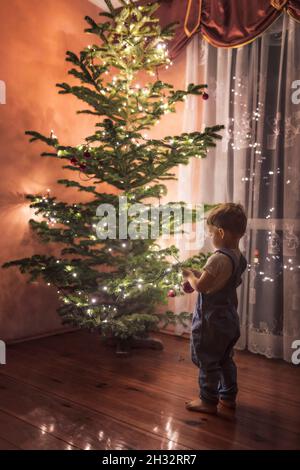 Kleinkind in blauen Overalls Dekoration Christmass Baum zum ersten Mal Stockfoto