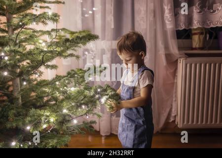 Kleinkind in blauen Overalls Dekoration Christmass Baum mit Ornamenten Stockfoto