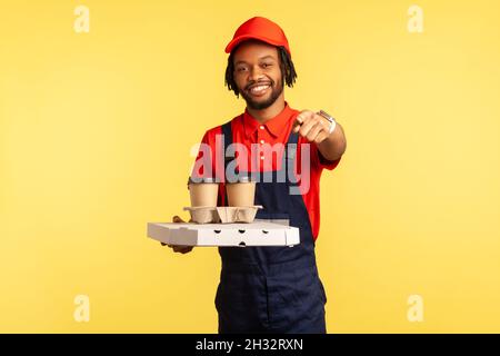Glücklicher lächelnder Lieferer mit blauen Overalls, der Kaffee und Pizza in einem Karton hielt und auf die Kamera und den Lieferauftrag zeigte. Innenaufnahme des Studios isoliert auf gelbem Hintergrund. Stockfoto