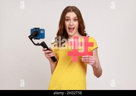 Aufgeregt Teenager-Mädchen mit Telefon auf Stabilisator führt Videoblog, Blick auf die Kamera mit überglücklich Ausdruck, zeigt rote Hashtag-Symbol. Innenaufnahme des Studios isoliert auf grauem Hintergrund. Stockfoto