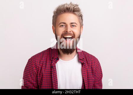 Porträt eines glücklichen, gutaussehenden bärtigen jungen Mannes im karierten Hemd, der mit offenem Mund aufrichtig lacht. Innenaufnahme im Studio, isoliert auf grauem Hintergrund Stockfoto