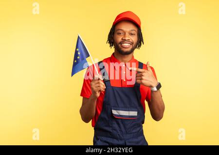 Fröhlicher Handwerker in Uniform mit Dreadlocks, die mit dem Finger auf die Flagge der Europäischen Union zeigen und mit positivem Ausdruck auf die Kamera blicken. Innenaufnahme des Studios isoliert auf gelbem Hintergrund. Stockfoto