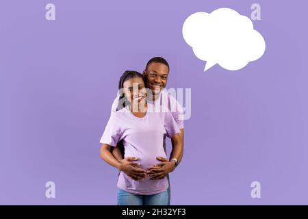 Porträt eines lächelnden schwarzen Mannes, der die Schwangeren von hinten umarmt, ein fröhliches Paar, das über den violetten Hintergrund des Studios steht und an das Baby-na denkt Stockfoto