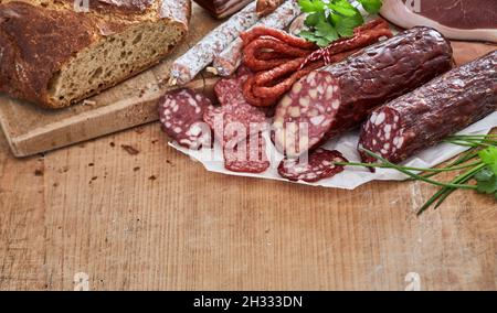 Schuss von trockengehärteten Würsten in der Nähe eines Laibs Brot auf einem Schneidebrett auf einem Holztisch mit Gemüse Stockfoto