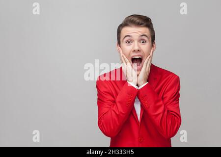 Oh mein gott. Portrait eines extrem staunenden Gentleman mit stilvoller Frisur in rotem Smoking und Fliege, die Hände an den Wangen hält und mit schockierten Augen die Kamera anschaut. Innenaufnahme im Studio, grauer Hintergrund Stockfoto