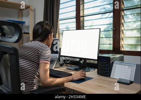 Freiberufliche junge asiatische Frau arbeitet mit leerem Monitor aus Laptop und Aufbewahrungsbox, Kalender auf Holztisch im Haus Stockfoto