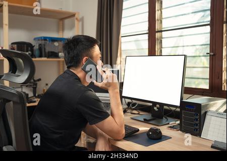 Freiberuflicher junger asiatischer Mann, der mit Handy und leerem Display telefoniert, Aufbewahrungsbox am Holzschreibtisch an der Fensterseite im Haus Stockfoto