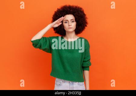 Ja, Sir. Strikte Frau mit Afro-Frisur in grünem Pullover, die mit Respekt und Verantwortung grütet, patriotische Bürger hören mit Ehre. Innenaufnahme des Studios isoliert auf orangefarbenem Hintergrund. Stockfoto