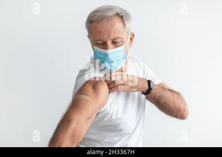 Covid-19-Schutz. Geimpfter Älterer Mann Mit Gesichtsmaske Zeigt Arm Mit Medizinischem Gips Nach Der Impfung Auf Grauem Studiohintergrund Sitzend. Ich gehe Stockfoto