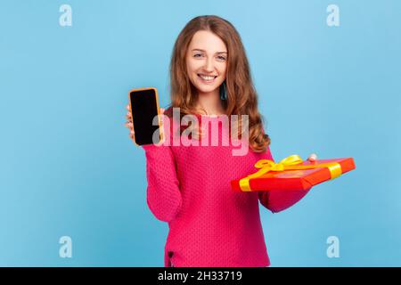 Weihnachtsgeschenk, Bonus für mobile Benutzer. Frau trägt einen rosa Pullover, hält eine Geschenkbox und ein Mobiltelefon mit leerem Display für Online-Shopping-Werbung. Innenaufnahme des Studios isoliert auf blauem Hintergrund. Stockfoto