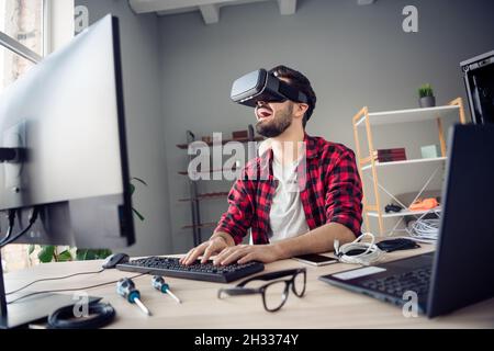 Portrait von attraktiven fröhlichen Kerl trägt Brille Headset spielen Videospiel Spaß am Arbeitsplatz Station drinnen Stockfoto