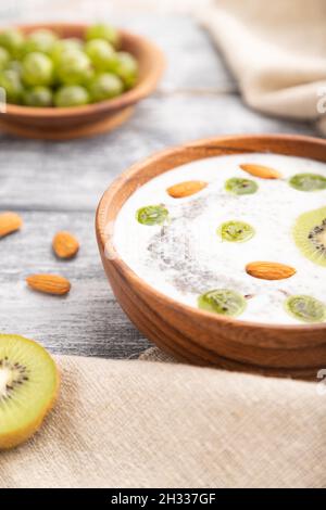 Joghurt mit Kiwi, Stachelbeere, Chia und Mandeln in Holzschüssel auf grauem Holzhintergrund und Leinentextilien. Seitenansicht, Nahaufnahme, selektiver Fokus. Stockfoto