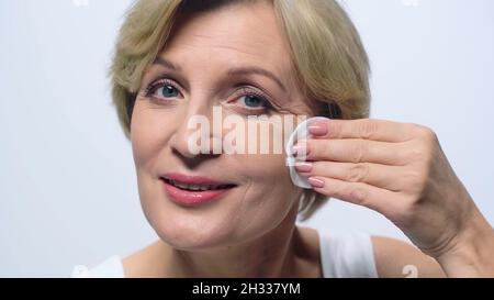 Positive Frau mittleren Alters wischende Gesicht mit Wattepad isoliert auf weiß Stockfoto