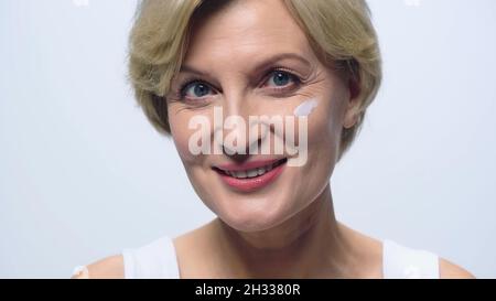 Fröhliche Frau mittleren Alters mit kosmetischer Creme auf dem Gesicht Blick auf die Kamera isoliert auf weiß Stockfoto