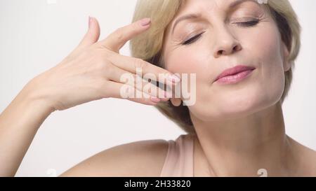 Nahaufnahme einer Frau mittleren Alters mit geschlossenen Augen, die das Gesicht isoliert auf Weiß berührt Stockfoto