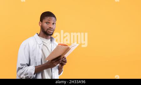 Emotionaler schwarzer männlicher Student, der Lehrbuch hält und vor Angst die Kamera anschaut, Probleme hat, auf gelbem Studiohintergrund, Panorama, für Tests zu studieren Stockfoto