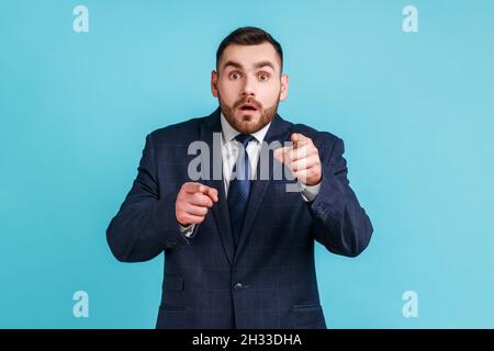 Erstaunt staunender, bärtiger Mann, der einen Anzug im offiziellen Stil trägt, der mit dem Finger auf die Kamera zeigt und mit vernehmlicher Ausdrucksweise auf die Kamera blickt. Innenaufnahme des Studios isoliert auf blauem Hintergrund. Stockfoto